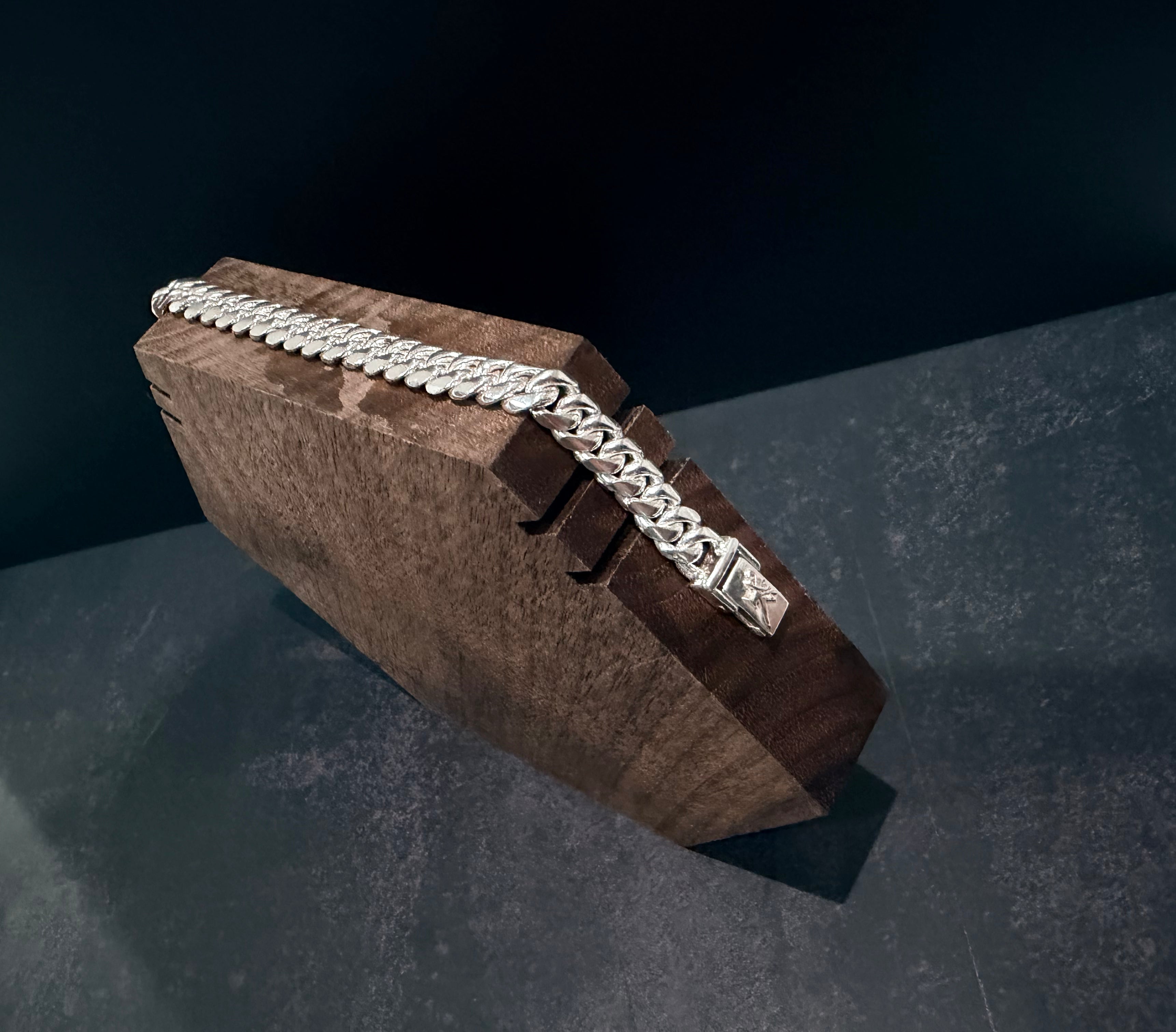 A silver-toned chain necklace draped across a dark brown wooden display block on a dark gray surface against a black background.