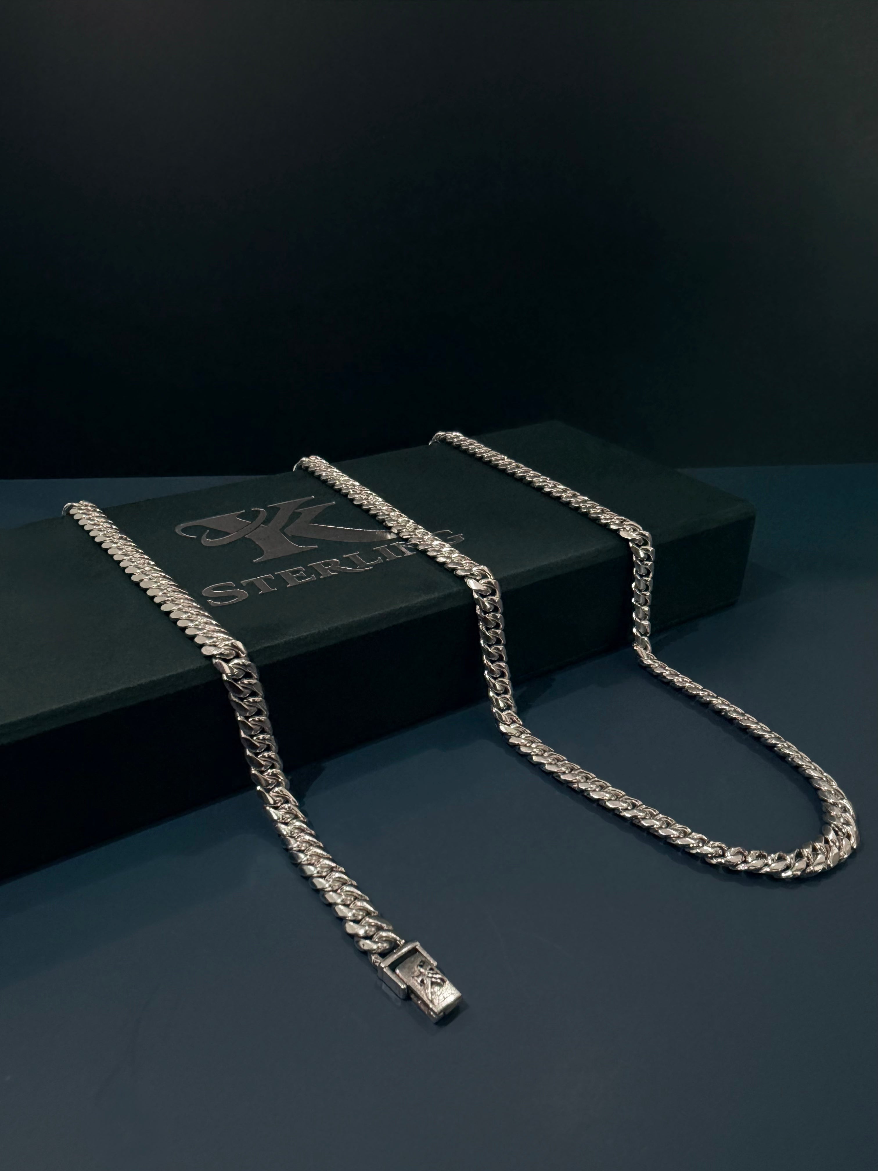 A silver-toned Cuban link chain necklace draped over a black jewelry box, photographed against a dark studio background.