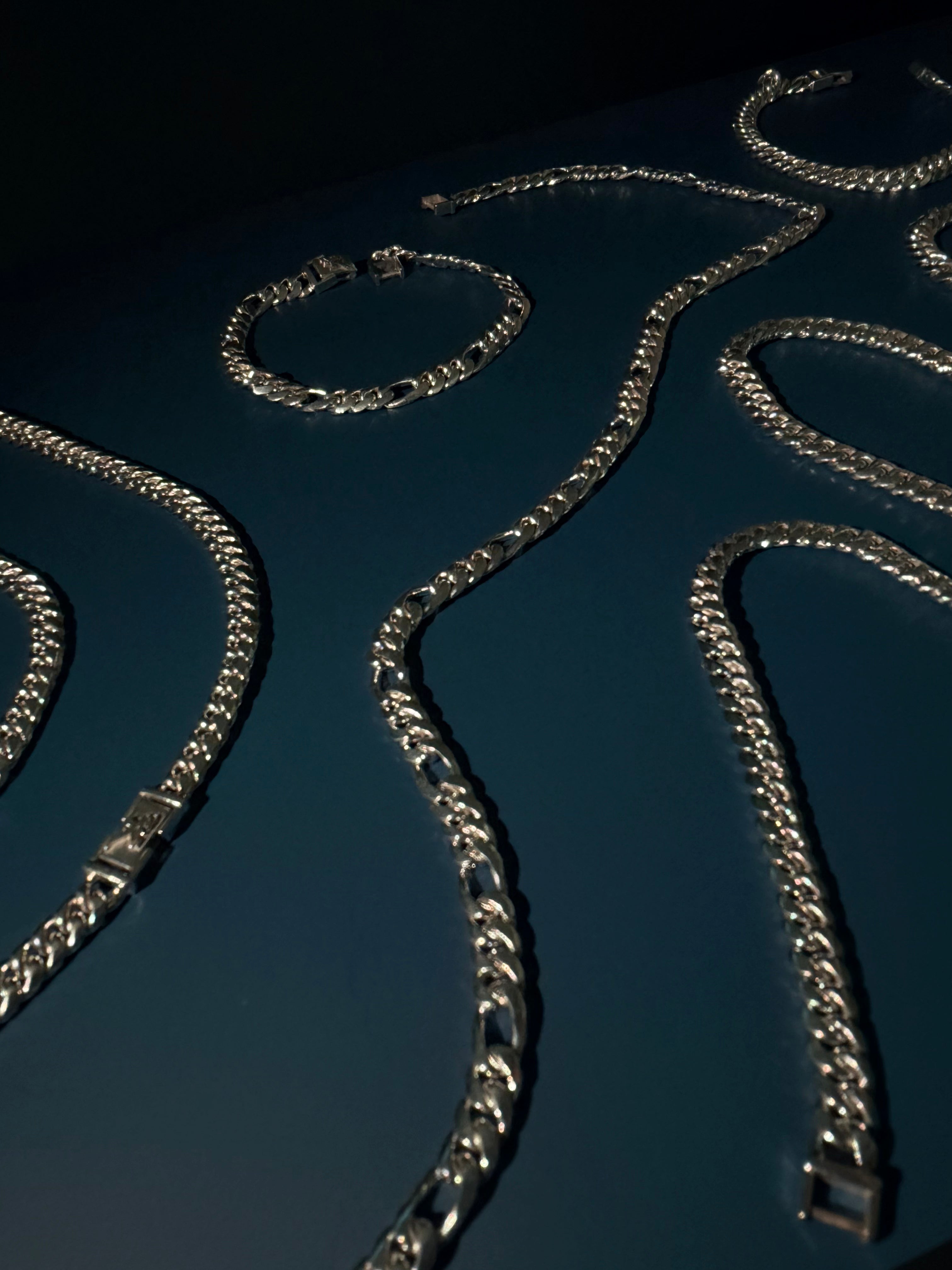 Several silver-toned curb chain necklaces and bracelets arranged on a dark blue surface under dramatic lighting.