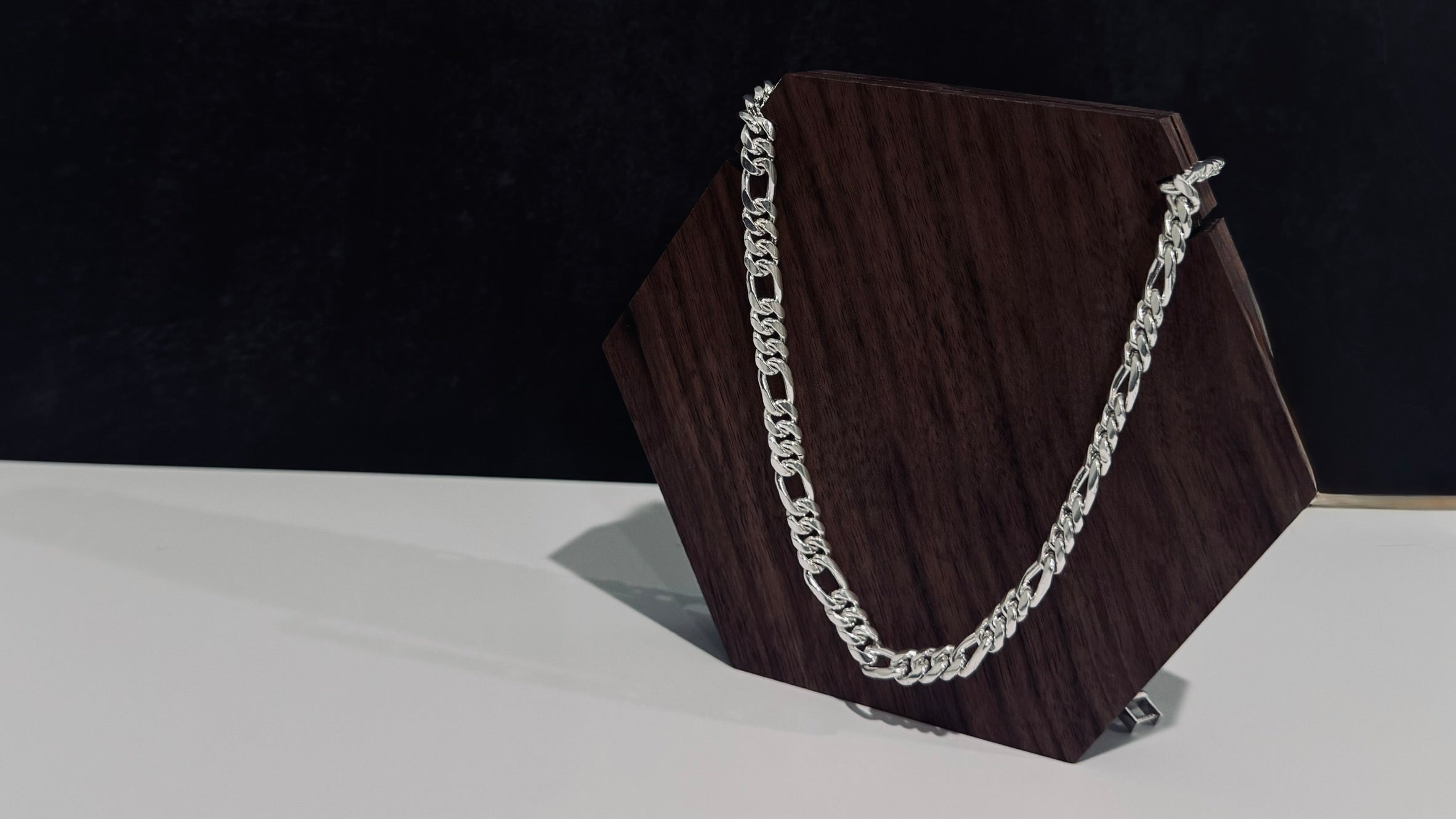 A silver-toned curb chain necklace draped over a dark brown hexagonal display stand against a black and white background.