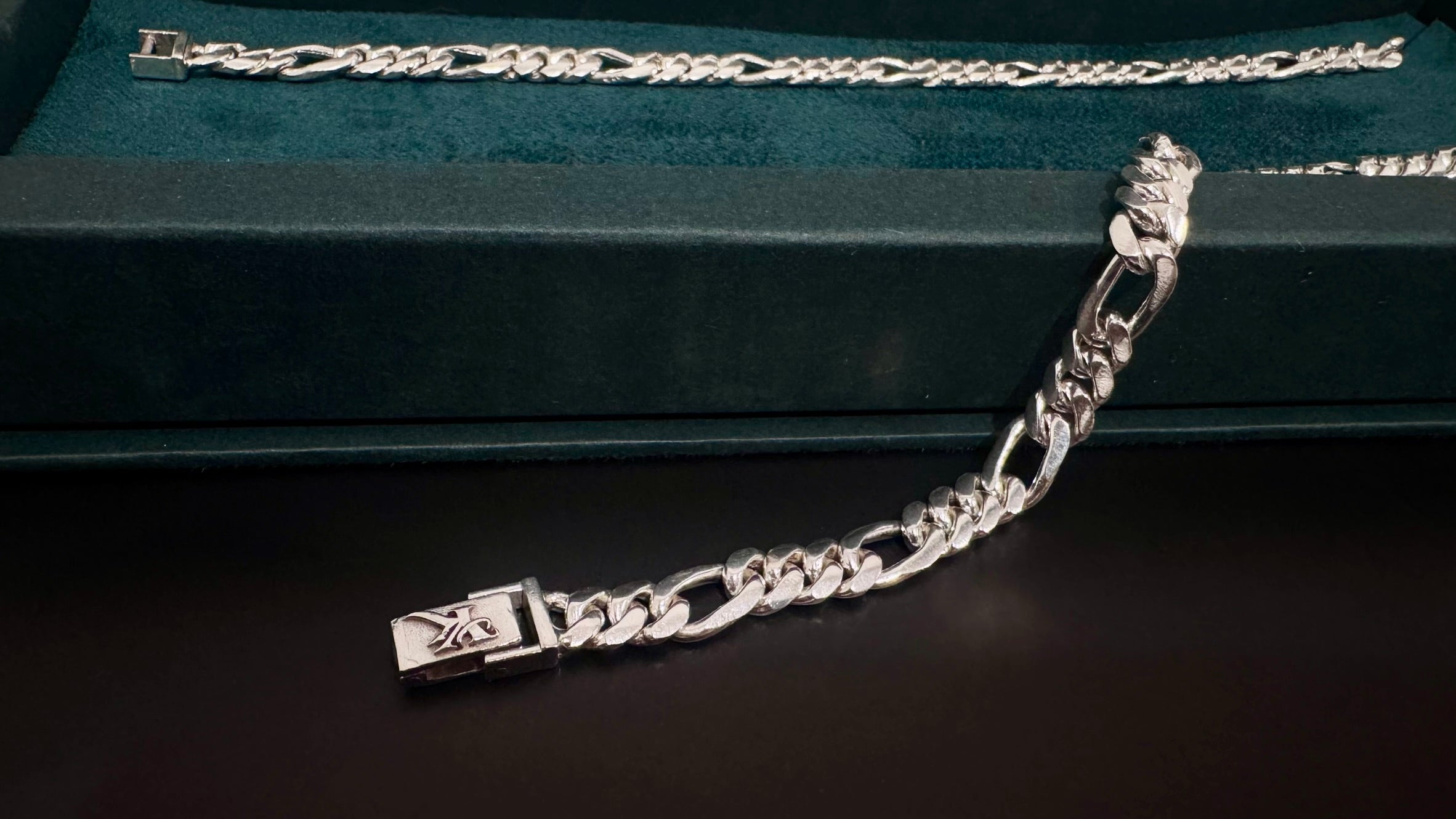 A silver-tone metal chain bracelet with a curb link design and a rectangular clasp, displayed on a dark green velvet jewelry box against a dark background.